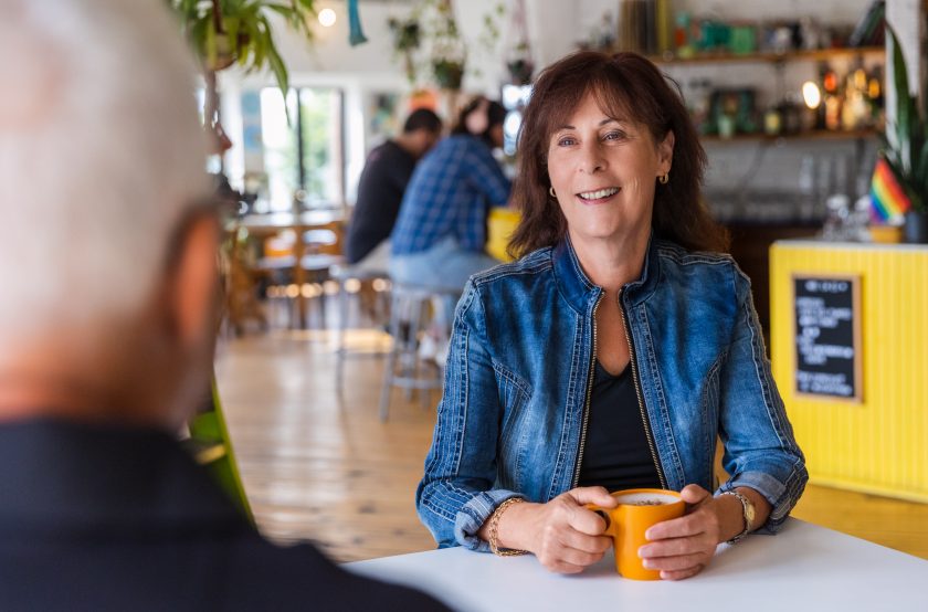 Femme assise à table dans un café avec une tasse à la main souriant à un homme assis en face d'elle.